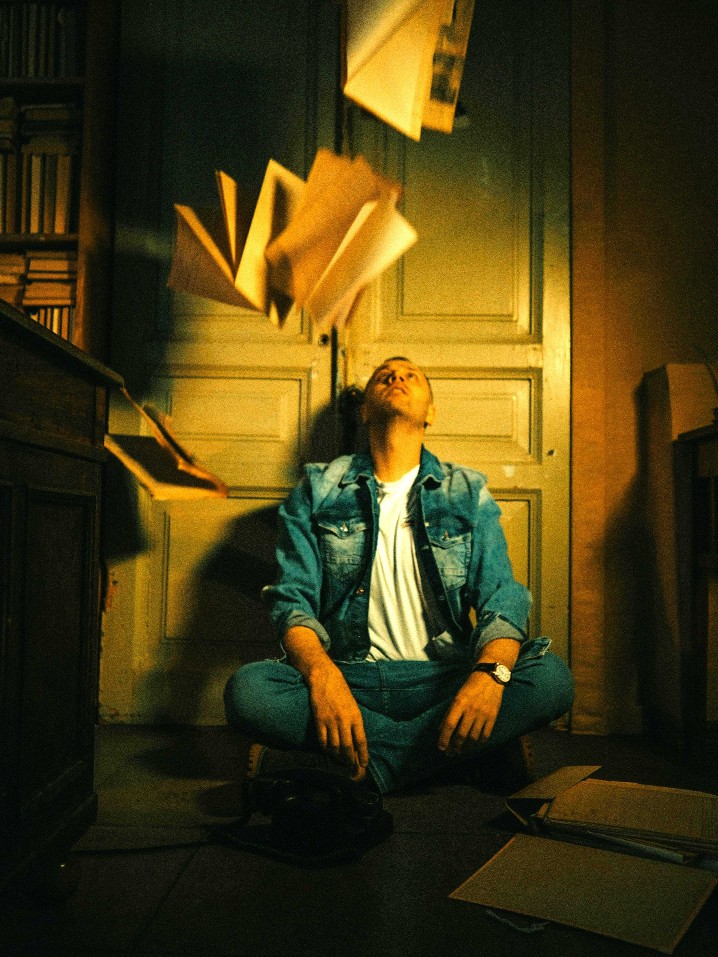 A young man sitting on the floor looks up at books thrown in the air - photo by Nadin Sh for Pexels